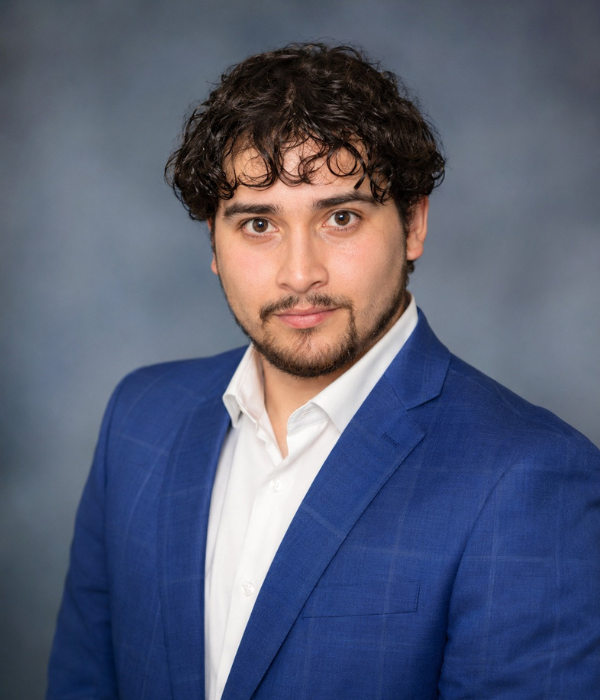 Professional headshot of Jovany Trevino wearing a blue blazer and white dress shirt, facing the camera with a slight smile against a softly blurred blue-gray studio background